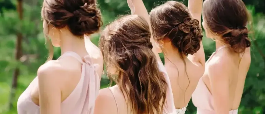 Štyri ženy v ružových šatách s rôznymi elegantnými účesmi na svadbu, zobrazené od chrbta na svadbe v exteriéri.