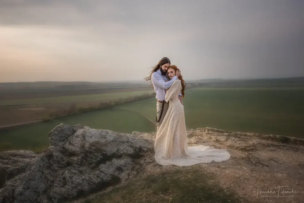 Mladomanželský pár oblečený v boho štýle pózuje na skale s výhľadom na polia.